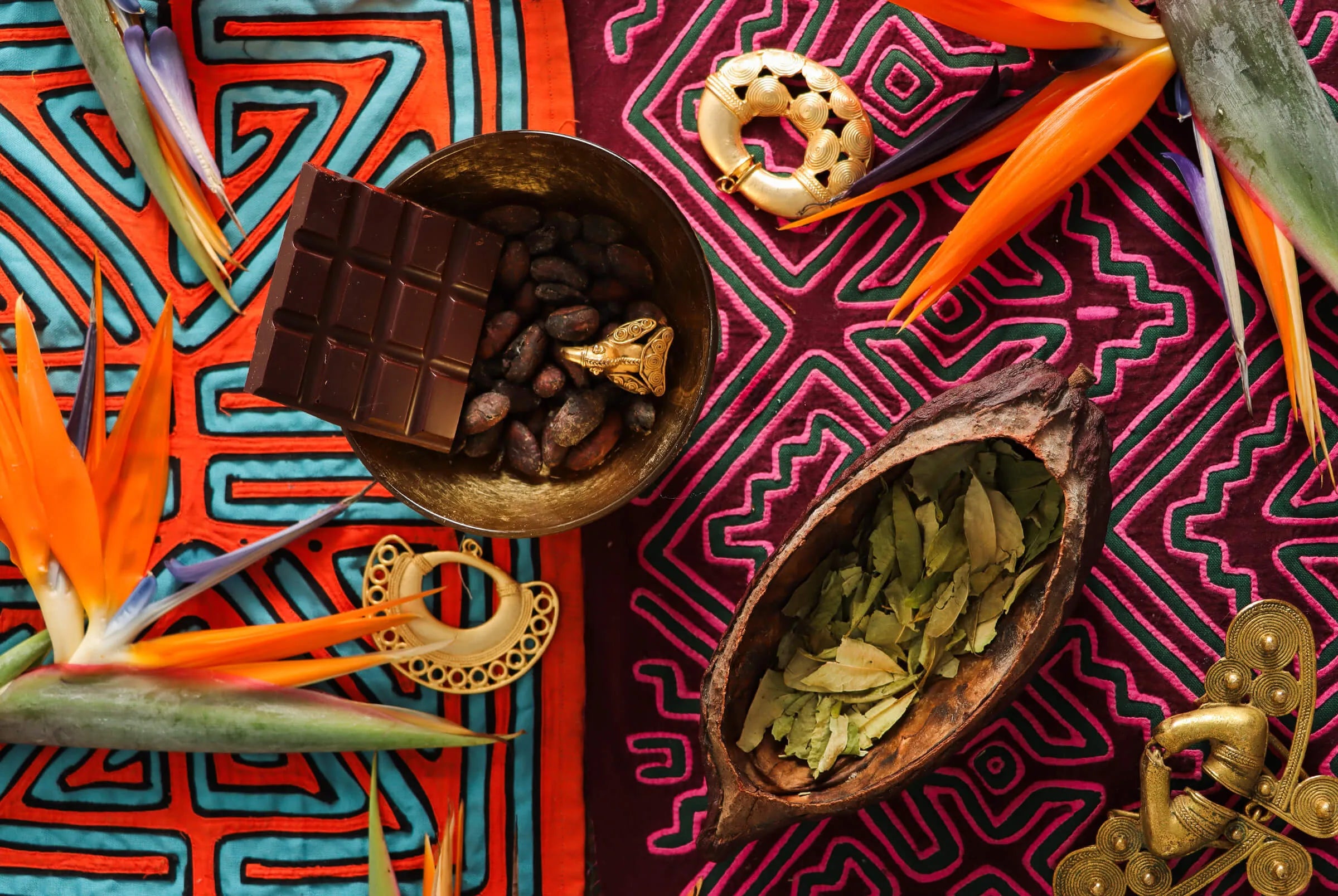 Cacao seeds, a piece of chocolate, dry coca leaves, golden artifacts, and tropical flowers arranged on colorful patterned fabric for a traditional cacao ceremony.