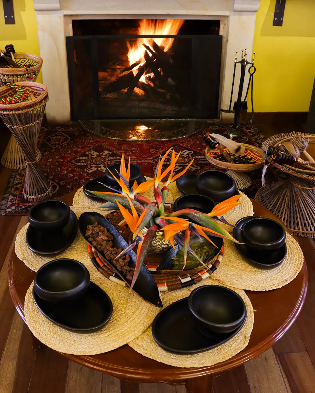 Set up for a cacao ceremony with a wooden table displaying black ceramic cups, a pitcher, pastries, and tropical flowers, in front of a lit fireplace.