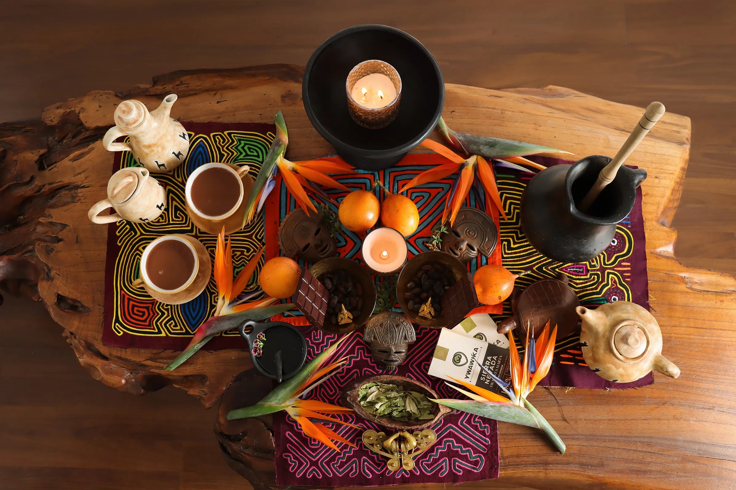 Set up for a cacao ceremony with a wooden table displaying tropical fruits and flowers, cacao seeds, dry coca leaves and golden artifacts over colorful patterned "mola" fabrics.