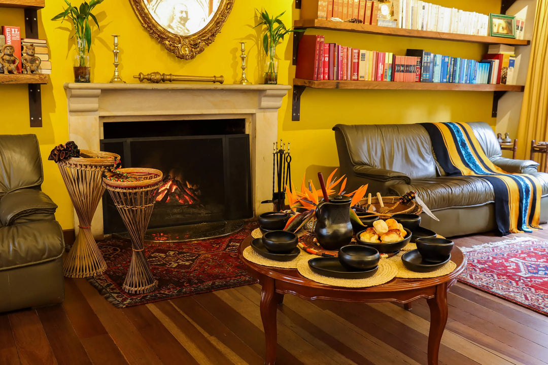 Cozy living room in a cultural heritage house in Bogotá, Colombia, set up for a cacao ceremony with a wooden table displaying black ceramic cups, a pitcher, pastries, and tropical flowers, in front of a lit fireplace and surrounded by bookshelves and leather sofas.